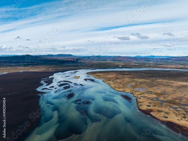 Obraz Wide aerial of a braided glacial river flowing across Icelandic plains under a blue sky, showing winding channels, sandbars, and contrasting land textures.