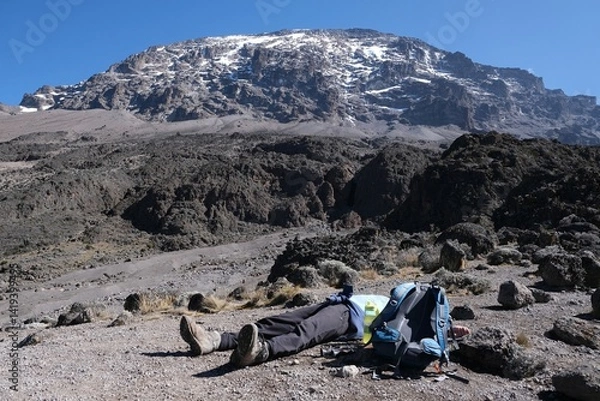 Fototapeta Silhouette of lying down exhausted man next ot backpack and Kilimanjaro peak in background, on trail from Barranco Hut to Barafu Camp, during trekking to Kilimanjaro Peak,  Machame Route. Tanzania