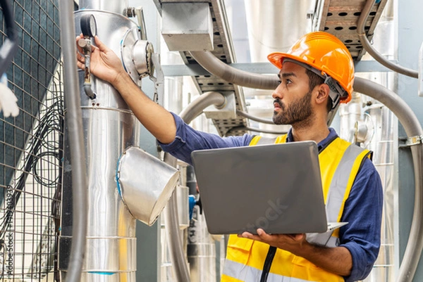 Obraz Male technical foreman in safety uniform inspects maintenance work work holding a laptop to look at plumbing and electrical systems on the roof of a building, Industrial background