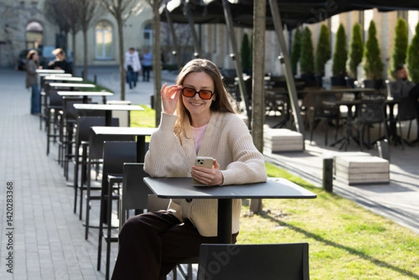 Fototapeta A young woman sits at an outdoor cafe table, smiling while looking at her phone. Sunlight illuminates the area, and empty tables are lined along the walkway