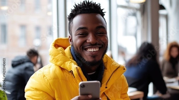 Fototapeta A smiling man with a beard stands in a modern office environment, wearing a tan jacket over a white shirt, holding a smartphone in his hands.