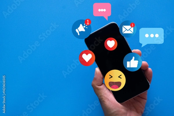 Obraz Hand holding smartphone with social media notifications, likes, hearts, and emoji icons on vibrant blue background. Concept of online interaction, social network, and digital communication.