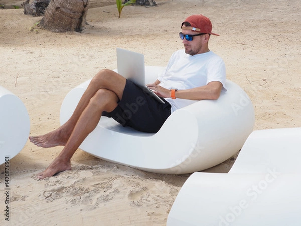 Fototapeta Man in a white t-shirt and sunglasses sits comfortably on white lounge chair at the beach remote working on laptop computer.