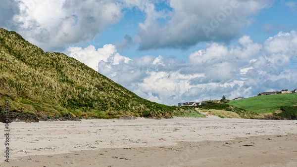 Obraz Idyllic coastal setting. A scenic beach backed by rolling green hills and charming houses. A calm atmosphere with a few people enjoying the day.