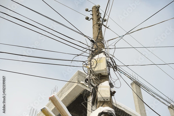 Fototapeta A weathered concrete utility pole overloaded with tangled electrical wires, network cables, and junction boxes against a blue sky, near a construction site.