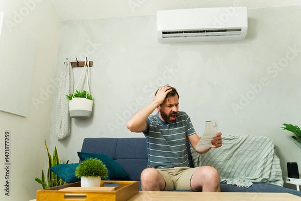 Fototapeta Worried man holding electricity bill sitting on sofa at home with air conditioning unit