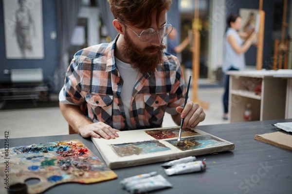 Fototapeta Man sitting by table and painting on small canvas
