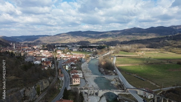 Fototapeta Breathtaking aerial view of the blue water Tanaro river flowing through the green hills of the countryside and the Roman bridge near Bagnasco city in the province of Cuneo