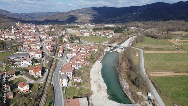 Fototapeta Breathtaking aerial view of the blue water Tanaro river flowing through the green hills of the countryside and the Roman bridge near Bagnasco city in the province of Cuneo