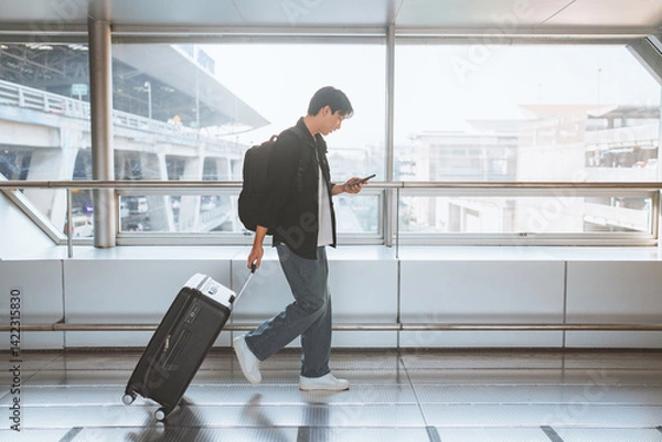 Fototapeta Side view - Male traveler walking with luggage using mobile smartphone at the modern airport terminal, man on way to flight boarding gate, Ready for travel or vacation journey, copy space