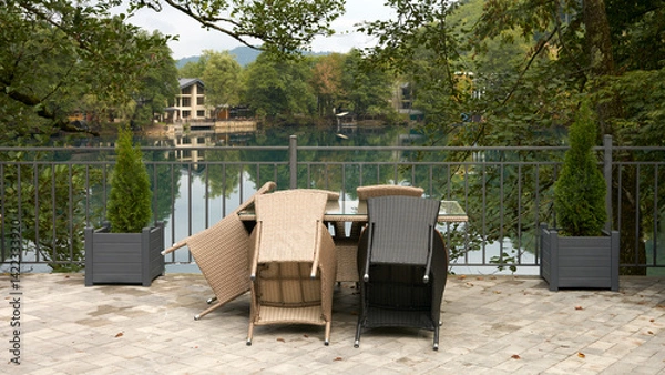 Fototapeta Panoramic view of a summer cafe on the shore of a picturesque mountain lake. A table with synthetic rattan chairs in the morning. Trees are reflected in the water.
