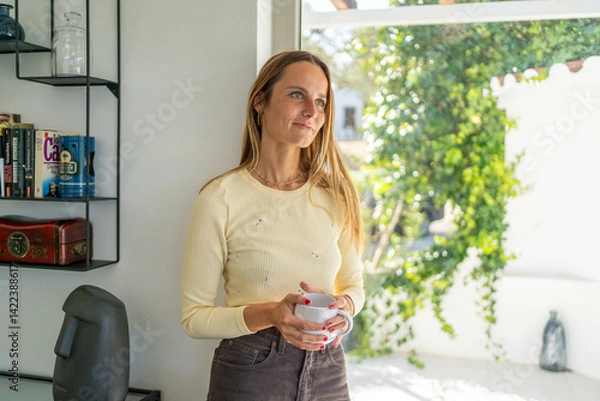 Obraz Woman by the window at home smiling, starting the day, with tea or coffee mug on her hand. Good morning, feeling calm and happy