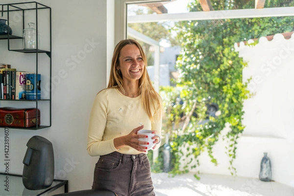 Obraz Woman at home by the window, starting new day, laughing with tea or coffee mug on her hand. Enjoying wellbeing, good morning, feeling happy, relaxing on weekend