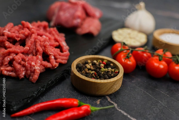 Obraz Close-up on a dark background in focus of peppercorns in a wooden bowl and shredded red minced meat with streaks of fat and red hot pepper surrounded by vegetables, meat and seasonings