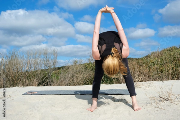Obraz Woman practicing yoga outdoors on sandy surface, performing forward bend with clasped hands behind back, embracing balance, flexibility, and mindfulness under bright sky and nature