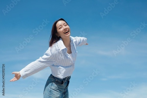 Obraz A cheerful Asian woman enjoying a sunny day at the beach. She is wearing a casual shirt and jeans standing in front of the blue sky background