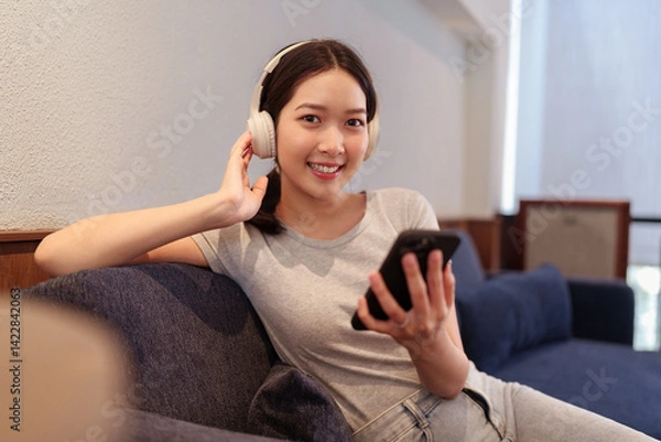 Obraz Young woman relaxing on sofa while listening to music with headphones and browsing songs on smartphone.