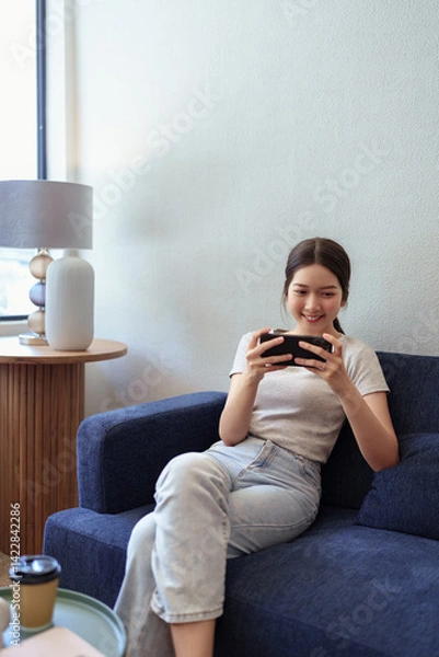 Obraz Young woman enjoying mobile gaming on smartphone while sitting on sofa in cozy daylight cafe corner