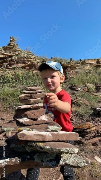 Fototapeta Little boy builds a pyramid of stones in the mountains