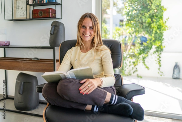 Obraz Woman reading a book, sitting in a chair, smiling at the camera, confortable, feeling happy, and relaxing on her free time