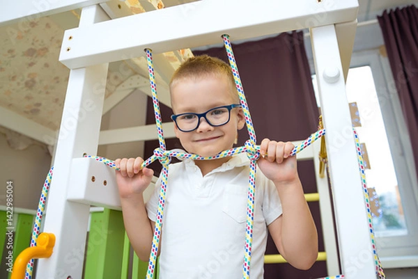 Fototapeta Boy performs gymnastic exercises on a wooden home sports complex with stairs and a slide. Sports exercises for children. Physical education of children at home
