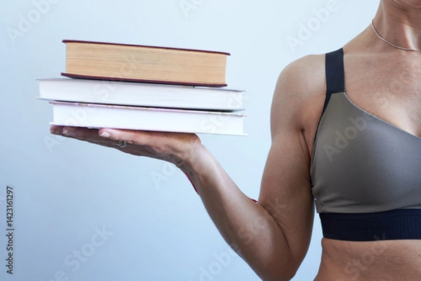 Fototapeta Woman in sports bra holding a stack of books on grey background. The concept is power in knowledge.