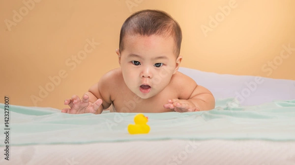 Fototapeta Asian baby lying on stomach with a bright smile, holding a yellow rubber duck on a mint green blanket. The joyful baby looks to the side against a warm beige background