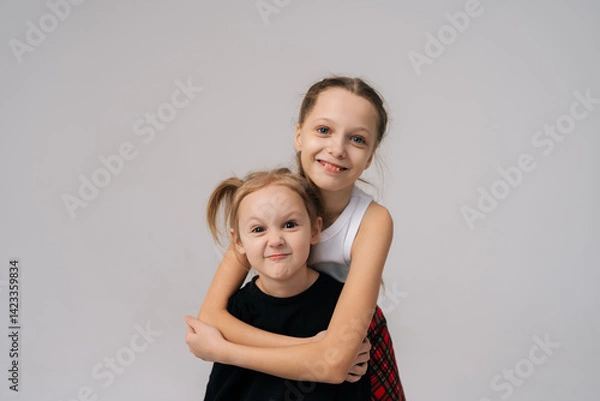 Obraz Adorable playful young sisters posing for studio portrait, with older sister affectionately hugging younger sister from behind, both smiling happily looking at camera. Concept of happy sisterhood.