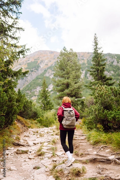 Fototapeta Woman traveler with backpack walking along rocky hiking trail in mountains. Happy woman enjoying nature and feeling freedom in open air. Hiking, adventure concept.