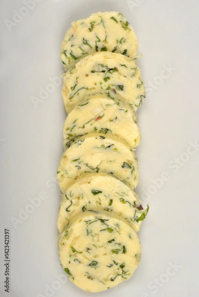 Fototapeta Slices of pale yellow butter with green herbs and garlic are arranged vertically on a white plate, captured from a top-down perspective