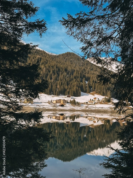 Fototapeta Calm mountain lake reflecting snowy landscape, houses and pine trees on a sunny winter day