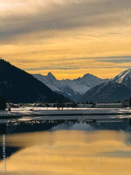 Fototapeta Calm lake reflecting golden sunset light, snow-covered buildings and mountains in the background
