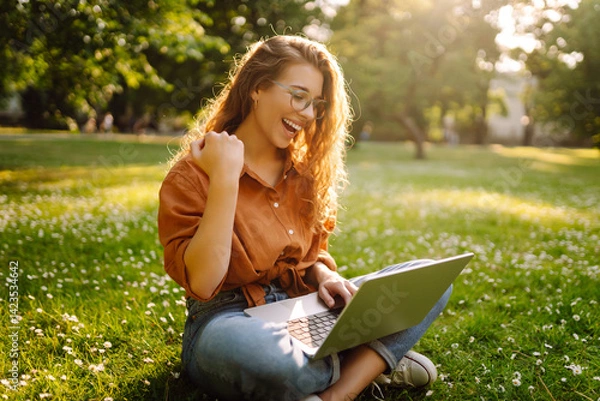 Fototapeta Young woman with laptop working on green lawn in park. Female freelancer with laptop enjoying sunny weather in park. Remote work concept, freelancer. Lifestyle.