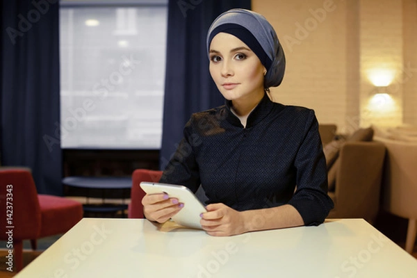 Fototapeta Beautiful Muslim woman in Islamic clothes sitting at a table with an electronic tablet in her hands