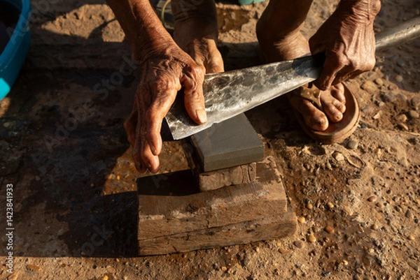 Obraz An elderly man works sharpening knives and machetes by hand, with a whetstone, at the Bai Don market in Pakse