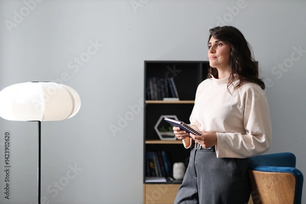 Fototapeta A woman is standing in front of a lamp and holding a tablet. She is wearing a white shirt and grey pants