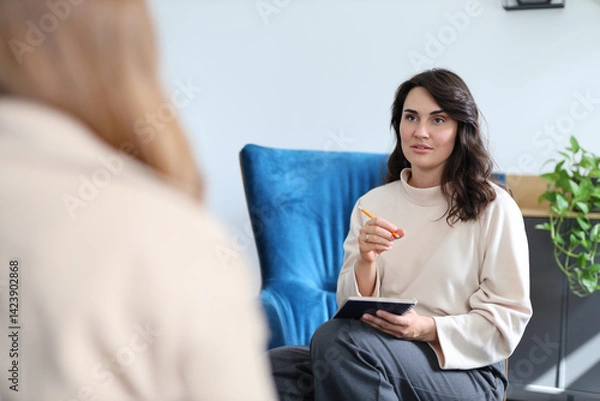 Obraz A woman is sitting on a blue chair with a notebook in her lap. She is looking at another woman who is sitting on a couch. The woman on the couch is holding a pen and a clipboard