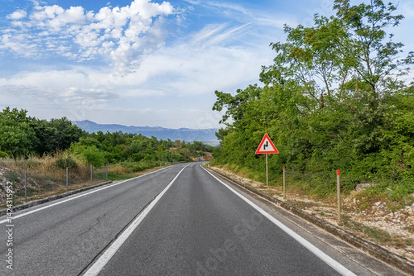 Obraz A winding country road stretches into the distance beneath a partly cloudy sky, with a traffic sign warning of upcoming curves surrounded by lush green foliage.