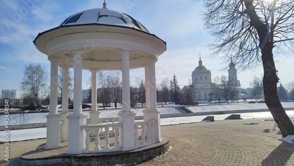 Fototapeta Rotunda on left bank of Orlik River with beautiful view of Cathedral of Archangel Michael in Orel city. Old white gazebo on background of river and Orthodox church on sunny winter day