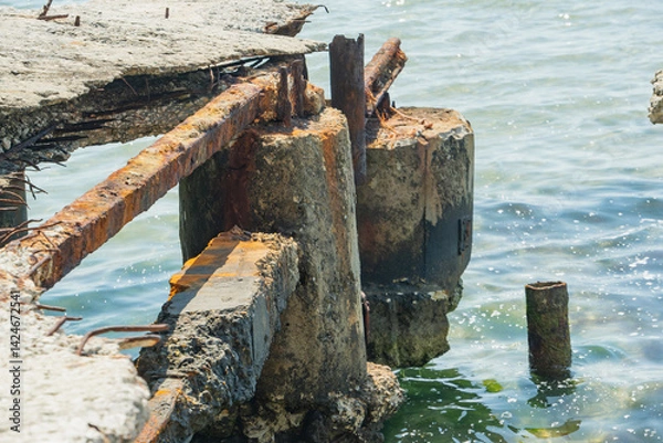 Fototapeta Close-up view shows the decaying remains of a concrete structure, with rusted metal and weathered surfaces extending into the sea
