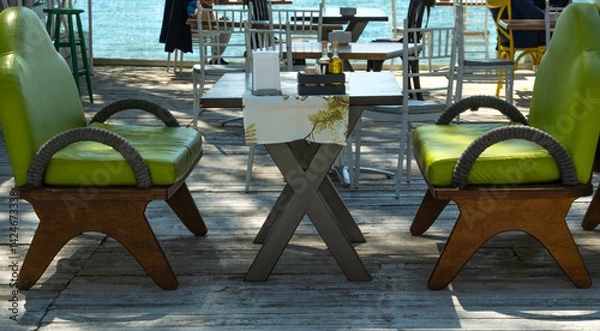 Fototapeta Outdoor dining area in a restaurant with two green chairs, table and sea view in the background
