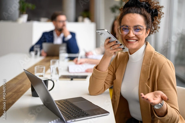 Fototapeta Happy manager in glasses speaking on her mobile phone's speaker while business coworkers collaborate in the background.