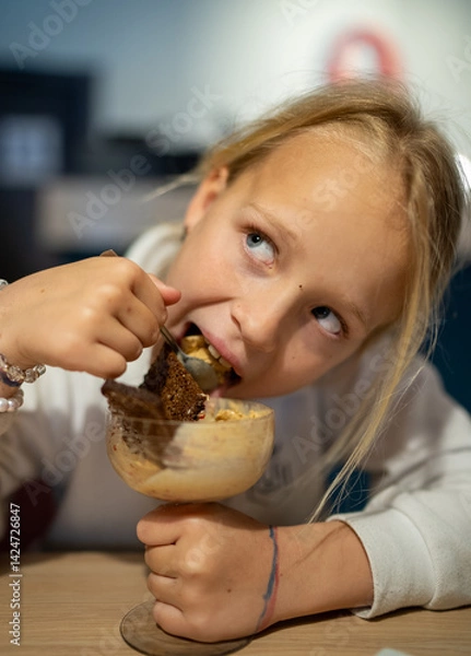 Fototapeta Blonde girl greedily eating ice cream with a slice of Borodino rye bread, holding a glass goblet, sitting at a table in a restaurant, looking away