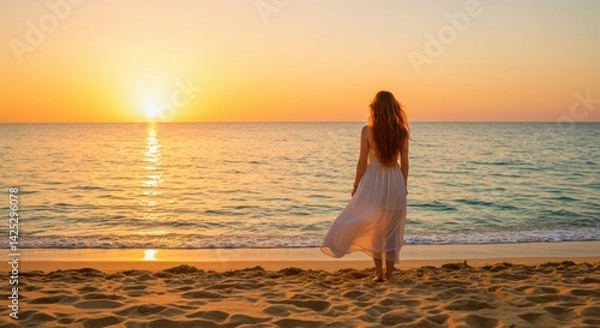 Obraz A woman stands alone on a quiet beach at sunset, facing the gently lapping waves. Her long hair flows in the soft ocean breeze, and she wears a flowing white dress that dances lightly with the wind