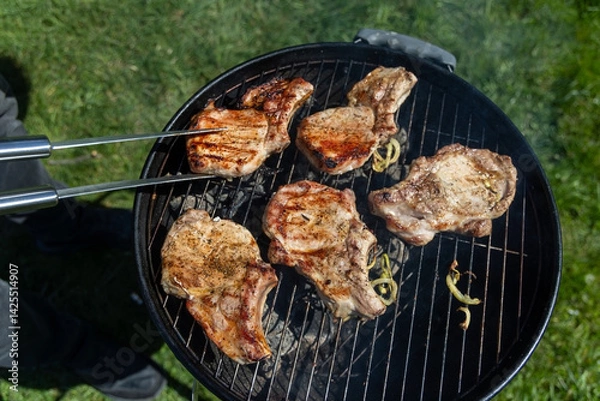 Fototapeta Grilled pork chops cooking on outdoor barbecue. Juicy, seasoned pork chops grilling over charcoal on a round barbecue grill during an outdoor cookout on a sunny day.