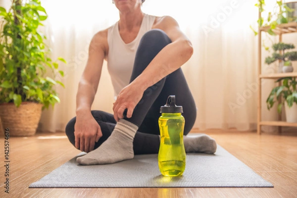 Fototapeta Mature woman taking a break from her fitness routine, sitting on a yoga mat with a water bottle, promoting healthy habits and wellness at home
