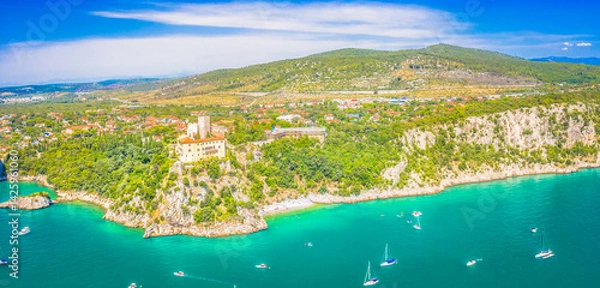 Fototapeta A wide panoramic view of the Adriatic coastline near Trieste, Italy, with the majestic landmark perched on a cliff, surrounded by lush greenery and overlooking the blue sea in a spectacular setting