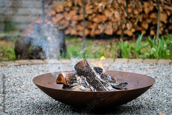 Obraz A close-up shot of burning firewood in a shallow, rusted steel fire pit placed on a gravel surface. Light smoke rises into the air while small flames dance around the blackened logs. 