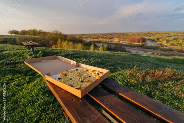 Obraz a pizza in a box stands on a bench on a hill with a beautiful view of the river, the concept of food in travel
