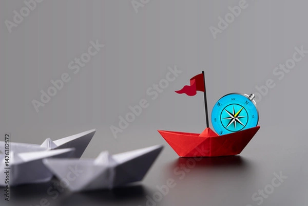 Obraz Red paper boat perched on a compass rose, surrounded by white boats, symbolizing direction and leadership.